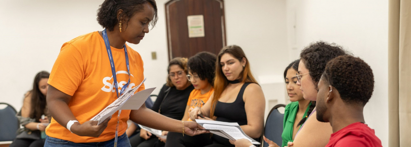 Jovens participando do curso de inglês no Sesc, recebendo materiais durante a aula.